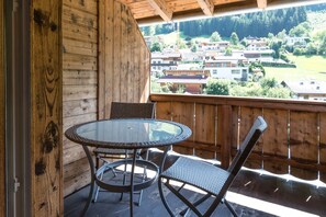 A wooden balcony with a glass table and wicker chairs, overlooking a mountainous village. - Hotel Wiesenhof (Mieders)