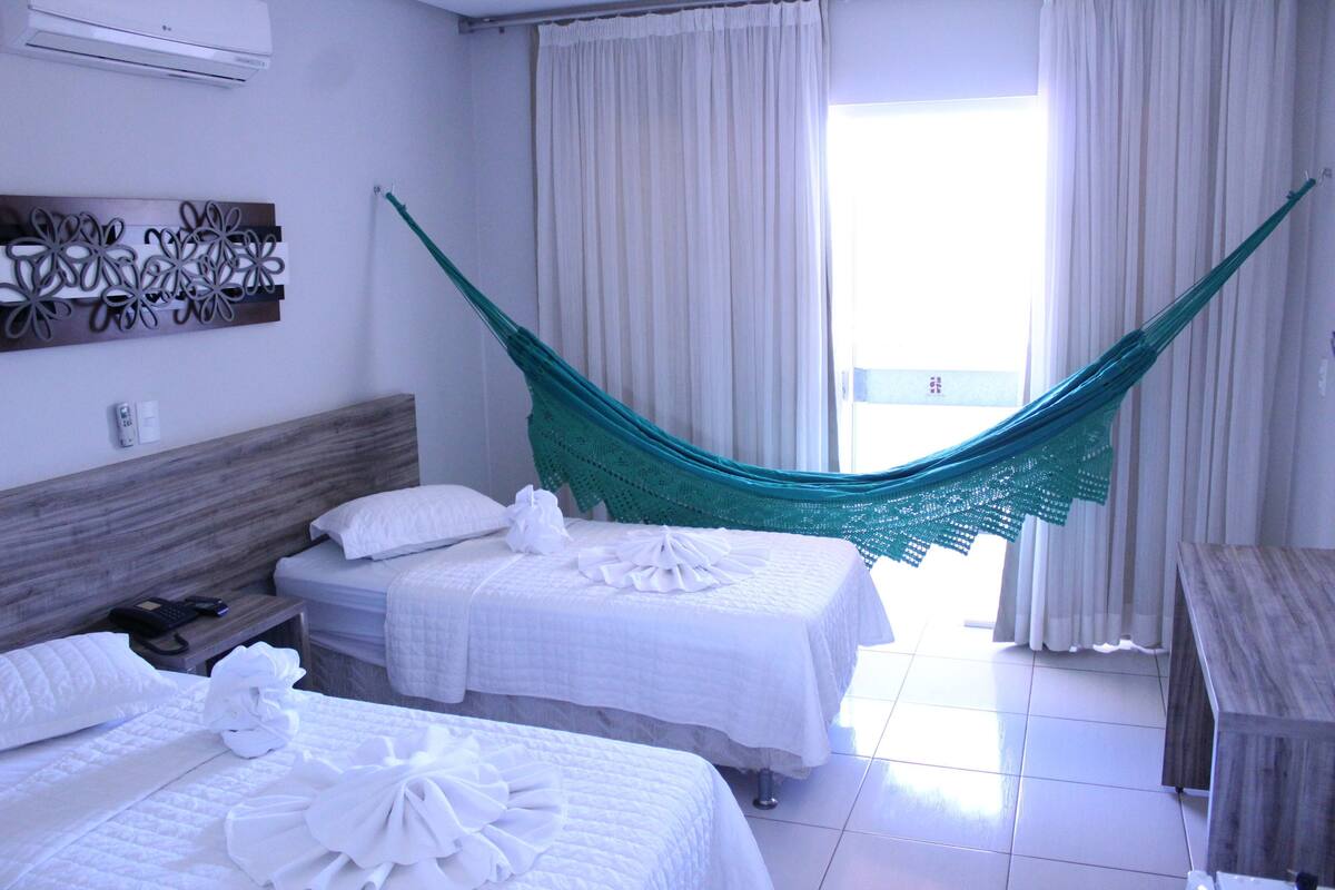 Hotelkamer met twee bedden, een hangmat, een houten hoofdbord, een wandairconditioner en uitzicht buiten door een raam.