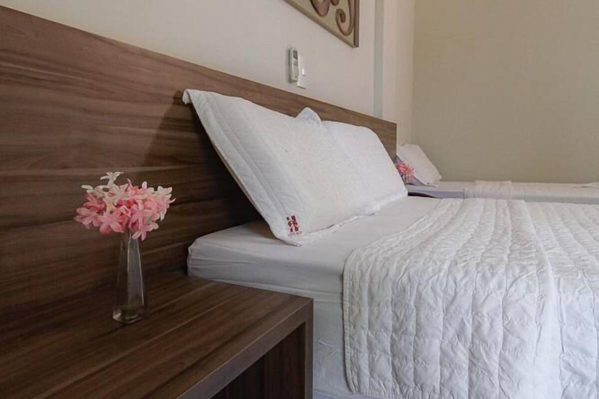 Een slaapkamer met een houten hoofdbord, een wit bed met een quilt, een nachtkastje met een bloemenvaas en een wandairconditioner.