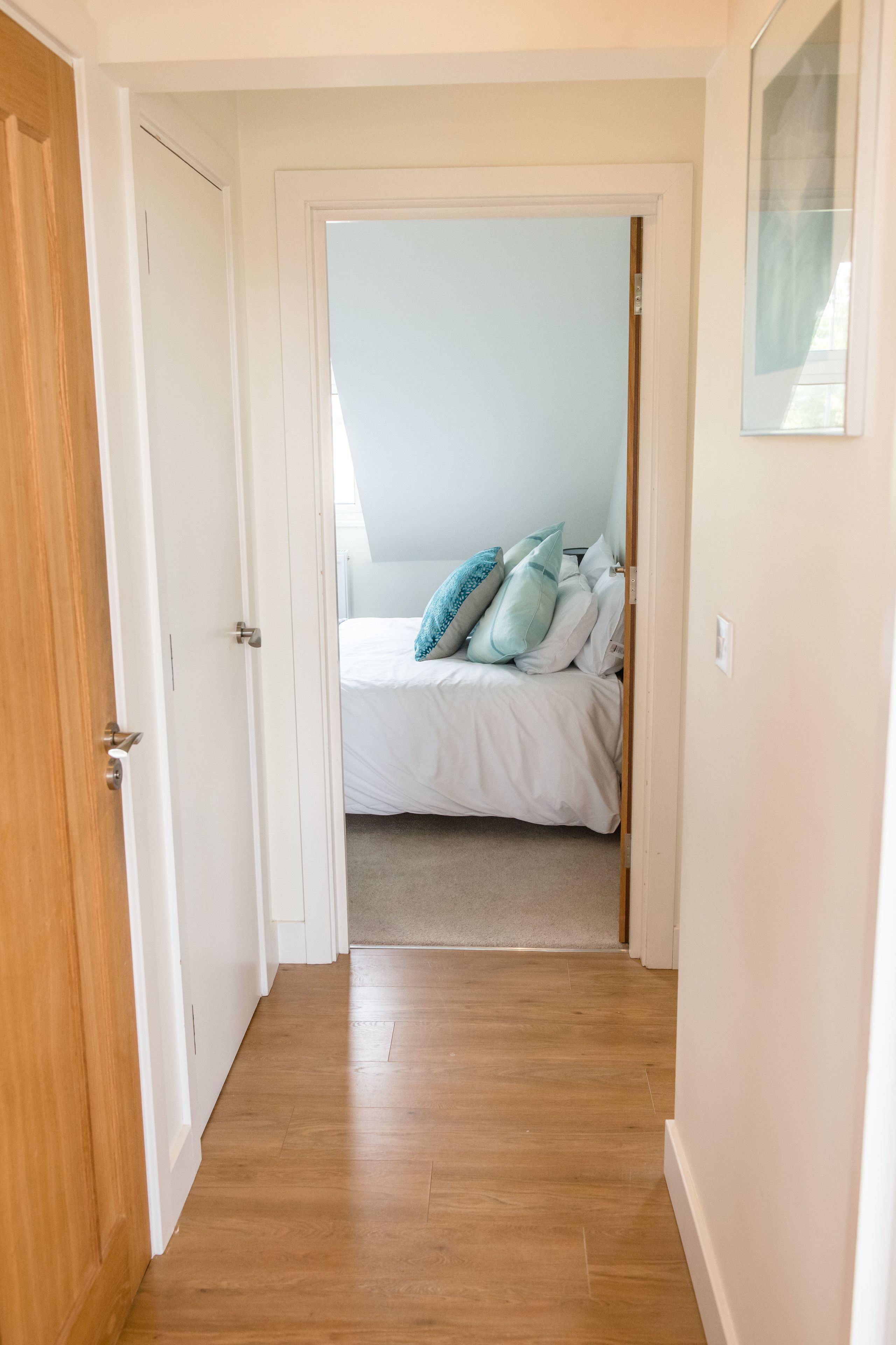 A narrow hallway leading to a bedroom with a bed, pillows, and a framed picture on the wall.