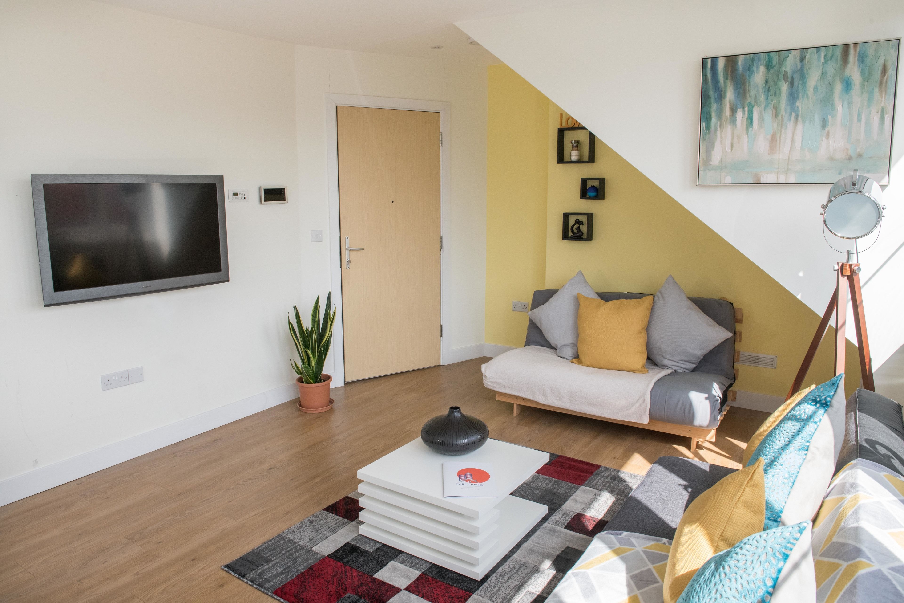A modern living room with a flat-screen TV, a sofa with cushions, a coffee table with books, a potted plant, and a painting on the wall.
