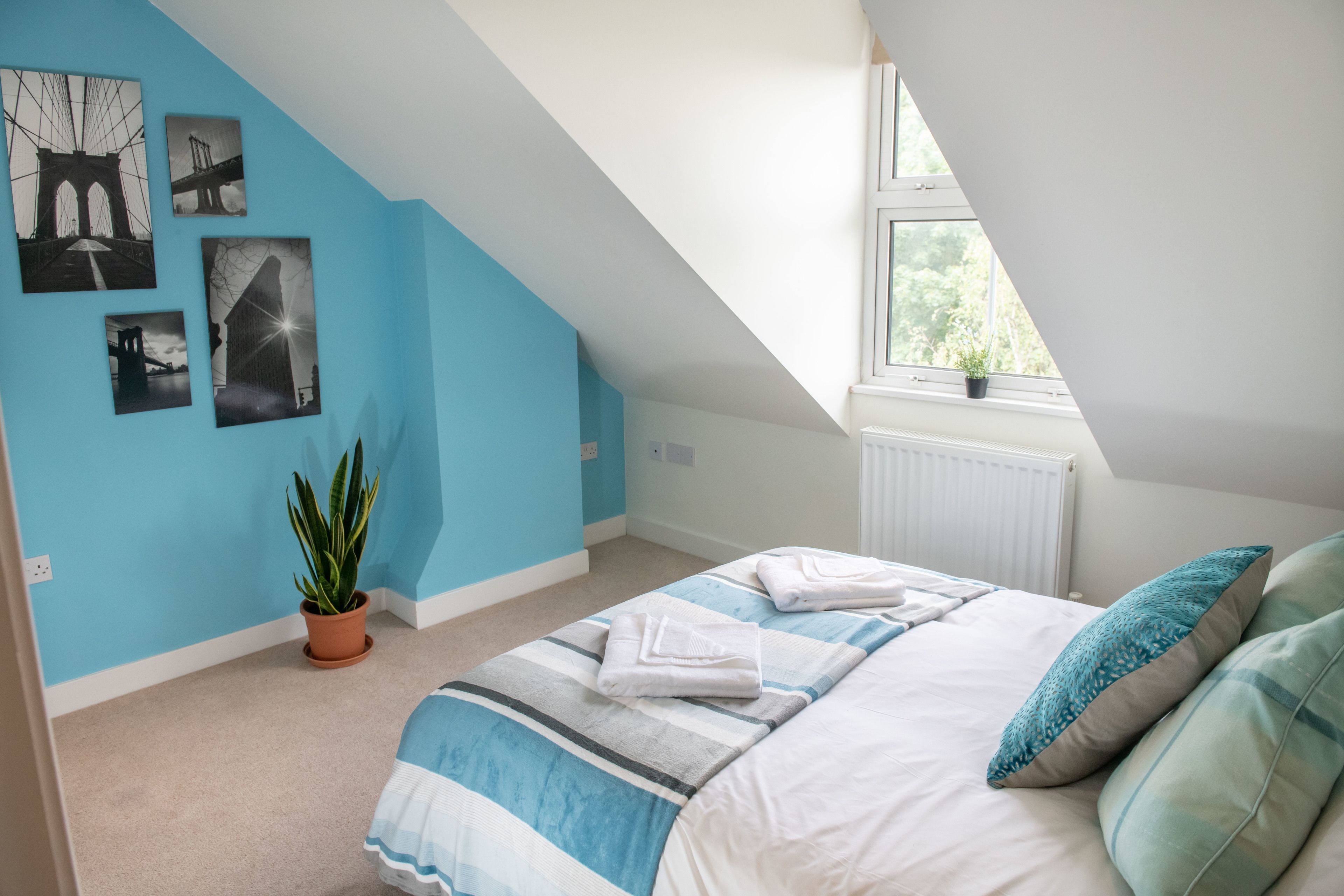 A bedroom with a sloped ceiling, a bed with blue and white bedding, a potted plant, and framed pictures on the wall.