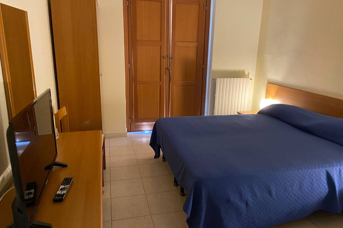 Une chambre à coucher comprenant un lit, un bureau avec un ordinateur et des télécommandes, une armoire et une porte.
