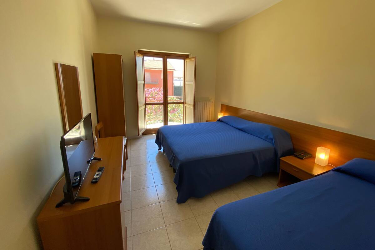 Une chambre d’hôtel avec deux lits, un bureau équipé d’un ordinateur et d’un téléphone, et une fenêtre donnant sur un bâtiment et des fleurs à l’extérieur.