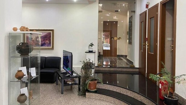 A lobby with a curved black floor, a glass display case with pottery, a black sofa, a television, and a wooden door. - Hotel Mercure (Castelluccio Inferiore)