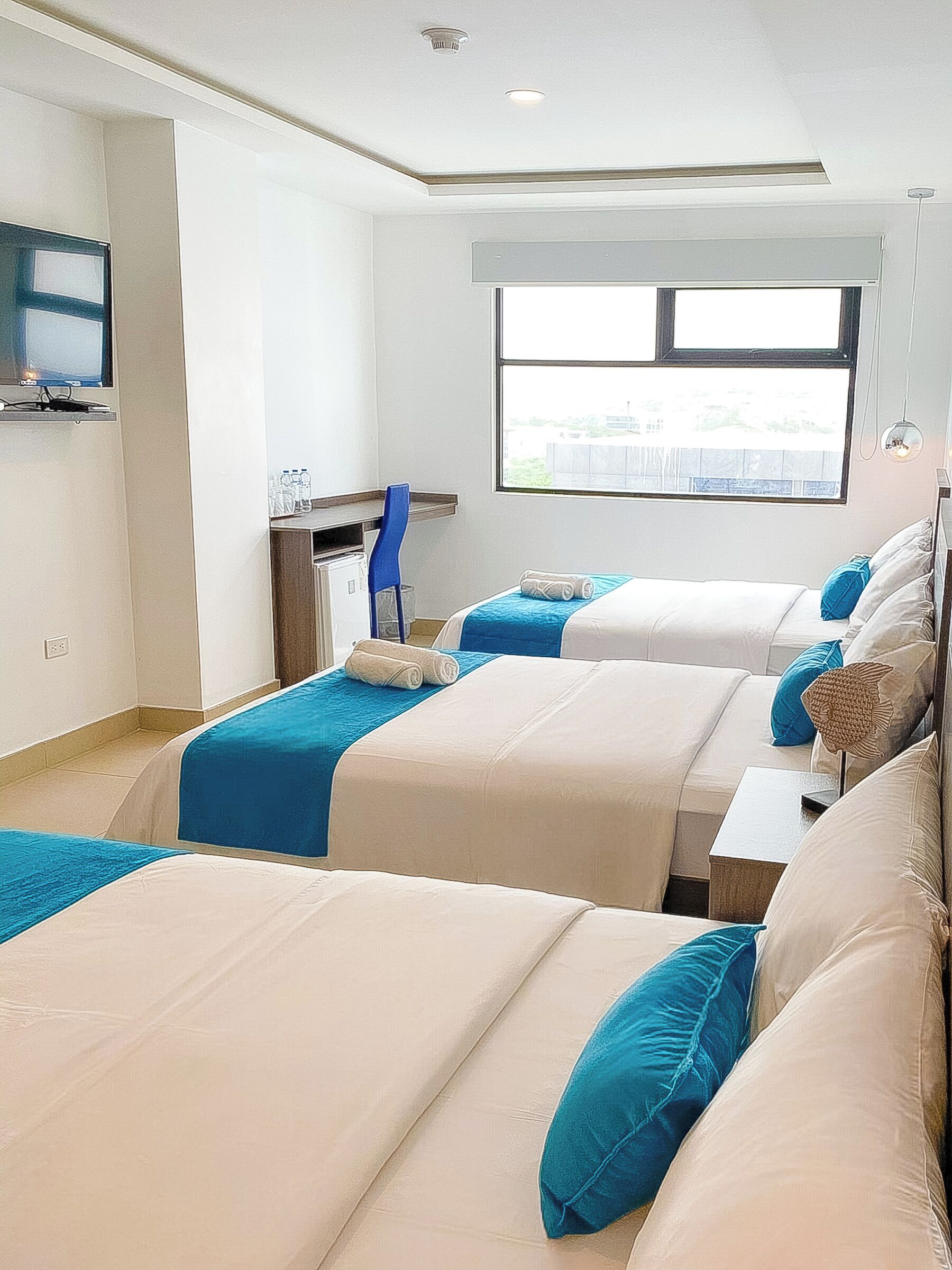 Habitación de hotel con tres camas, televisor, escritorio y ventana con vistas.