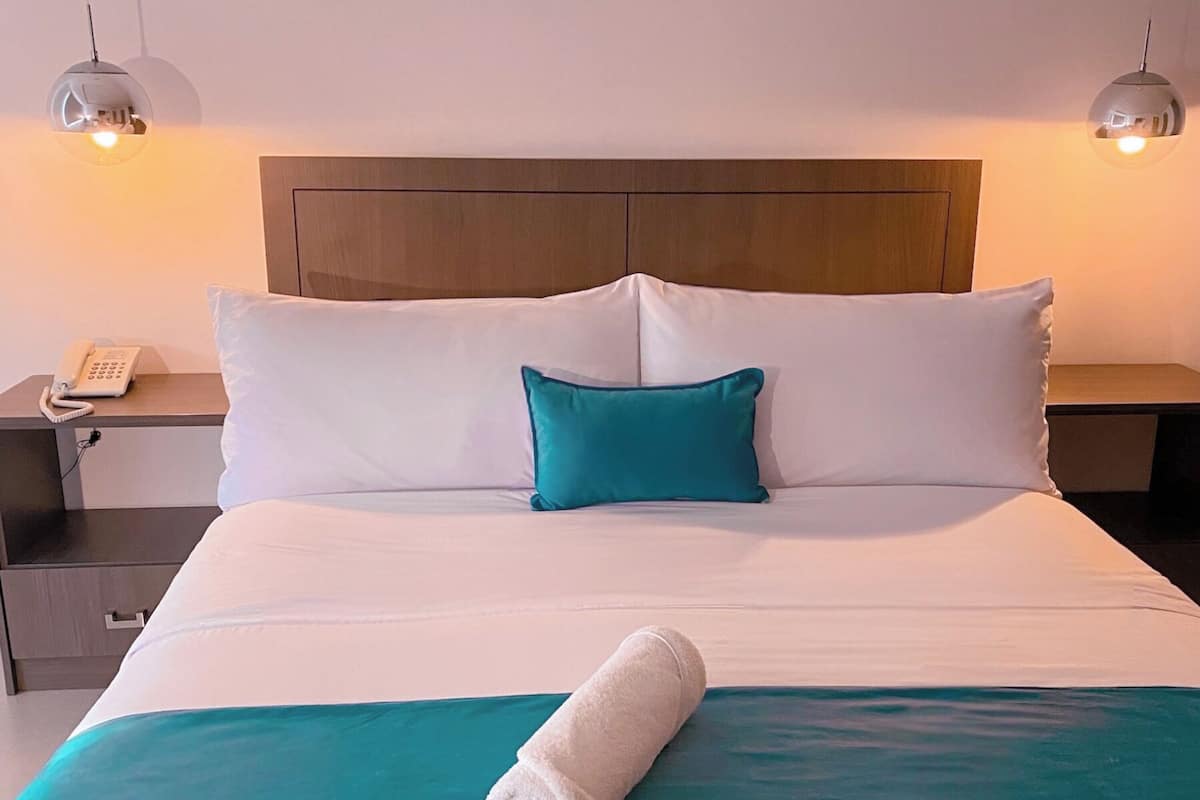 A neatly made bed with white and teal bedding, a teal pillow, and a rolled white towel on the bed. There are two bedside tables with lamps and a phone on one.