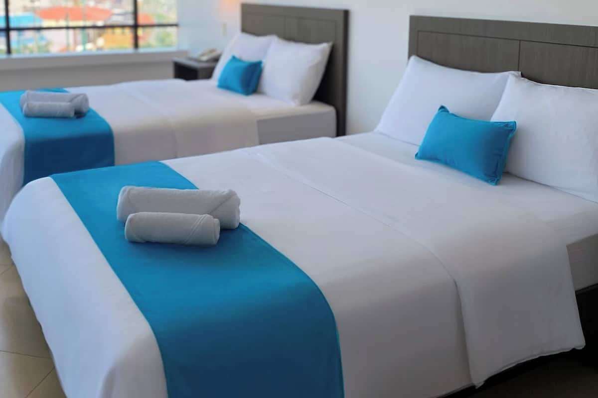 Two twin beds with white and blue bedding, a nightstand, a lamp, and a city view through large windows.
