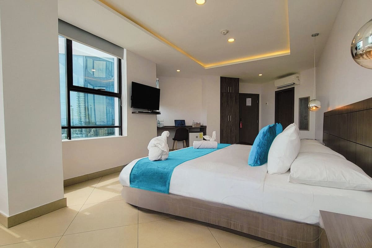 A modern hotel room with a large bed, a desk, a TV, and a view of the city through the window.