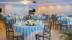 Un salón de banquetes con mesas redondas cubiertas con manteles azules, dispuestas en filas. Cada mesa tiene servilletas blancas, cristalería y centros de mesa florales. Las paredes muestran un letrero que dice "NAUTILUS EVENTS".
