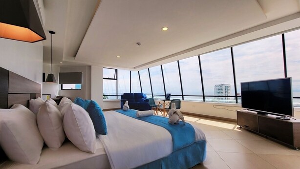 Una habitación de hotel moderna con una cama grande, un televisor y vistas a la ciudad a través de amplias ventanas.