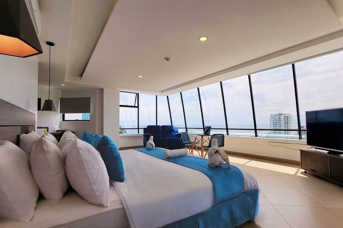 A modern hotel room with a large bed, a TV, and a view of the city through large windows.