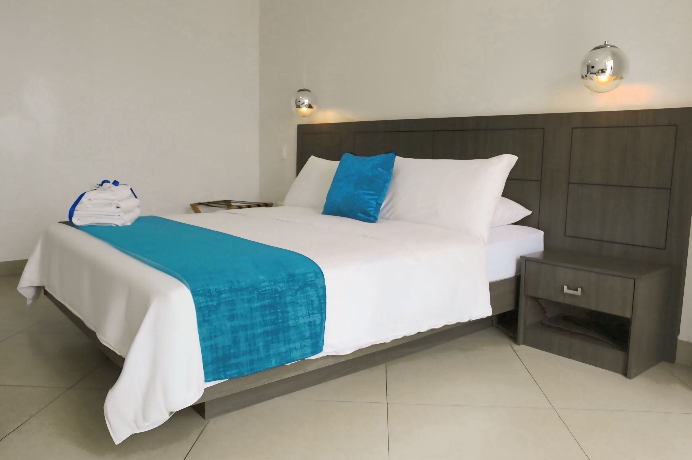 A neatly made bed with a blue blanket, a white pillow, and a blue accent pillow. There is a small bedside table with a lamp and a folded white bag on it.