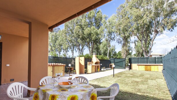 Un patio al aire libre con una mesa lista para comer, rodeado de sillas blancas, y vistas a un jardín trasero con zona de barbacoa y árboles.