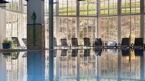 An indoor swimming pool with clear glass walls, chairs along the poolside, and a view of the outdoors. - Hotel Molitors Mühle (Eisenschmitt)