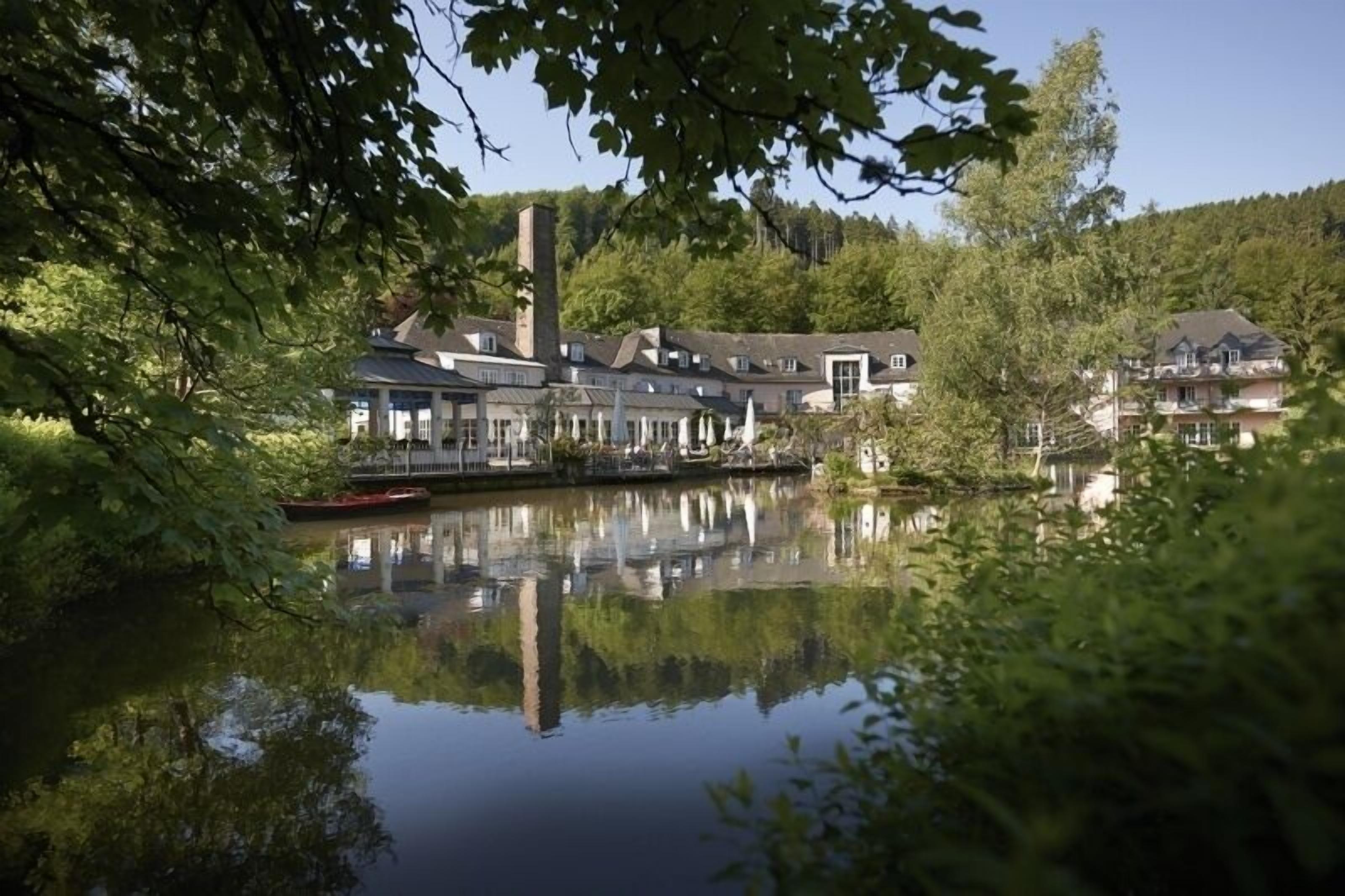 Un hôtel situé au bord d’un lac, doté d’une grande cheminée, dont la façade se reflète dans l’eau, et entouré d’une végétation luxuriante.