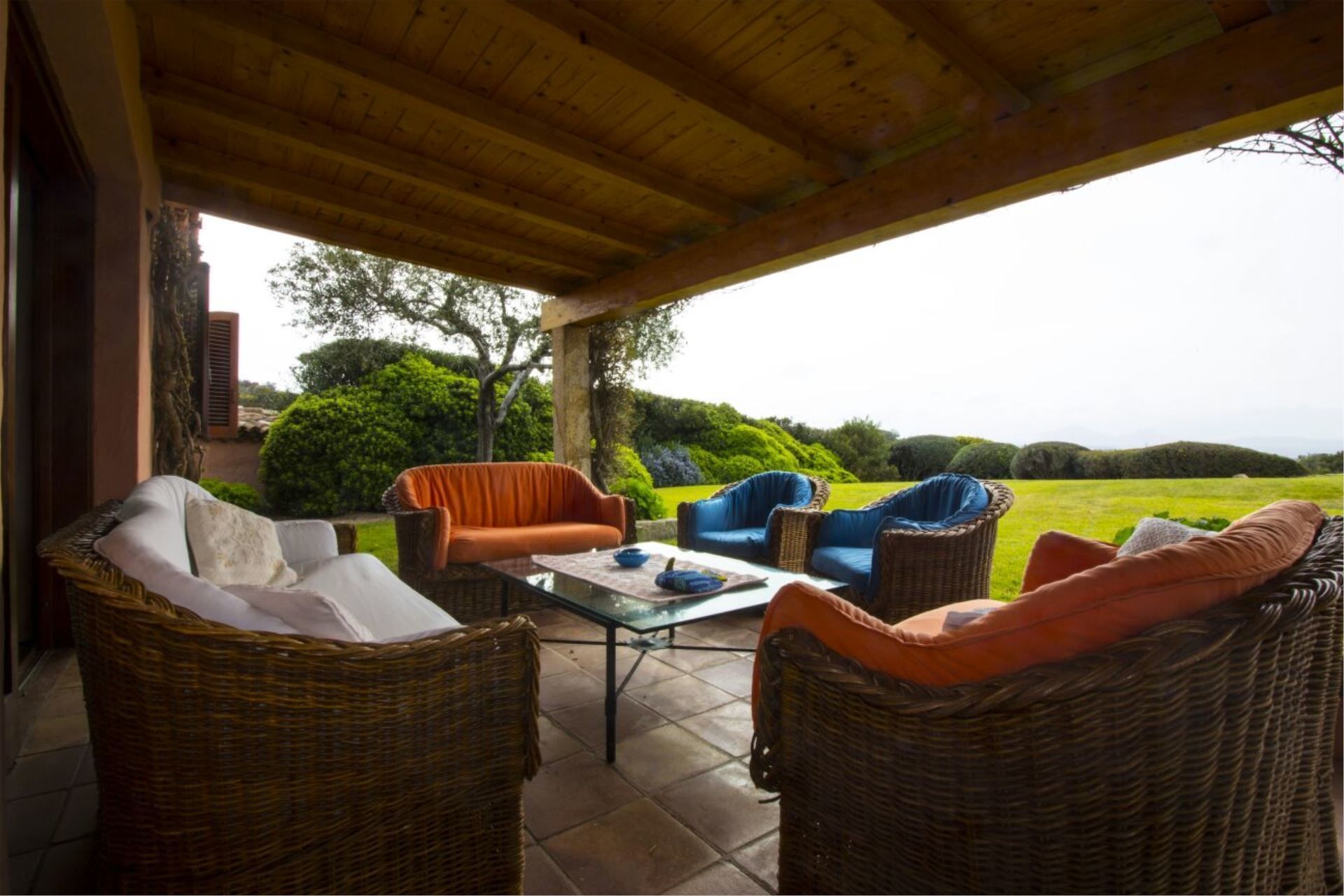 An outdoor patio with wicker furniture, a glass table, and a view of a green landscape.