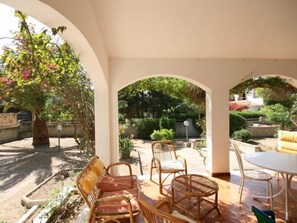 An outdoor patio area with wicker furniture, a round table, and a view of a garden with various plants and a tree. - Villa Eleonora (Maracalagonis)