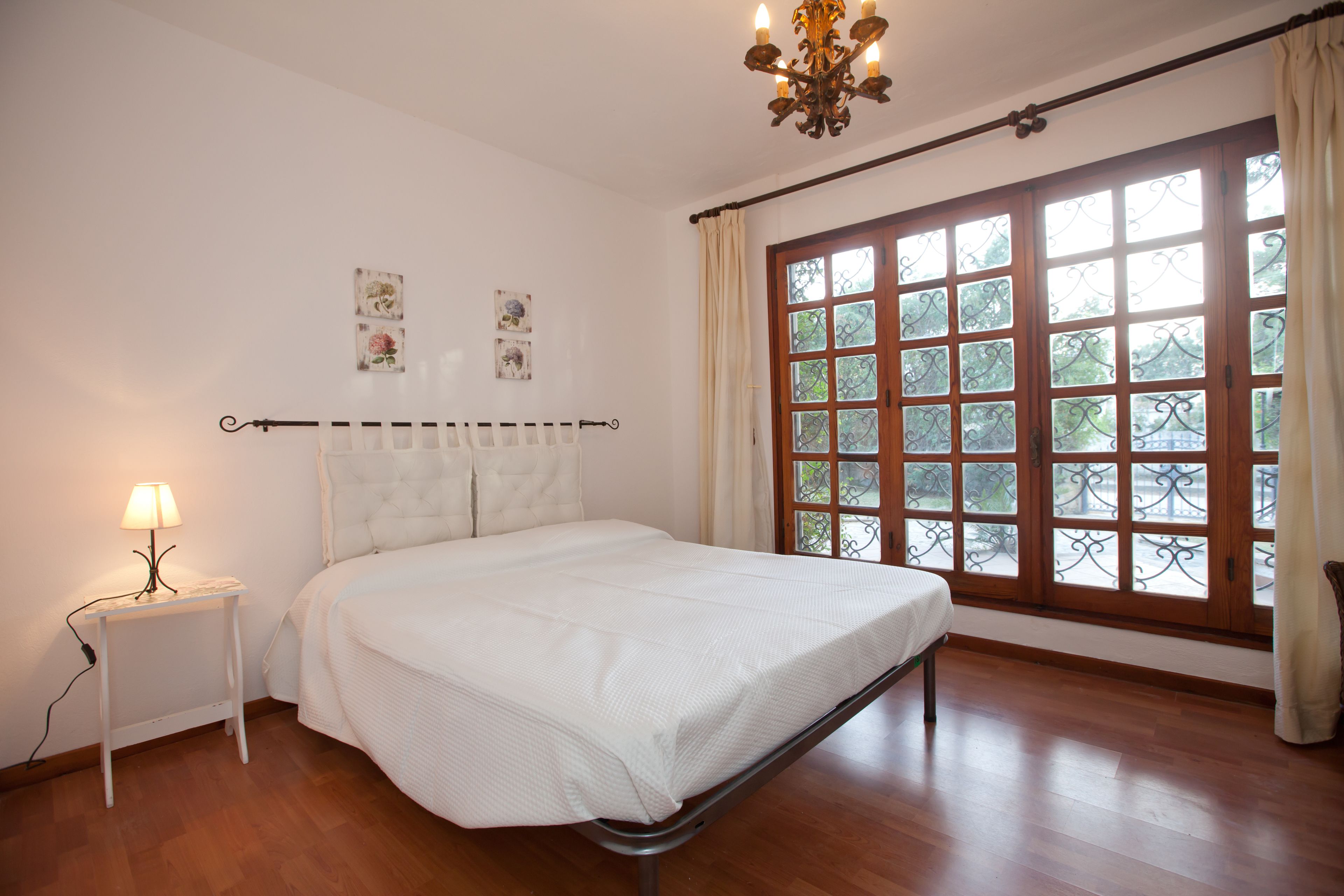 Un dormitorio con una cama, una mesita de noche con lámpara, un candelabro y un ventanal con cortinas.