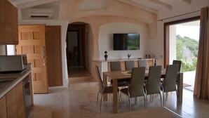 A modern kitchen with wooden cabinets, a dining area with a table and chairs, a TV mounted on the wall, and a view of greenery outside. - Villa La Mendula (Arzachena)