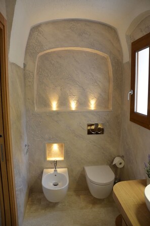 A compact bathroom with a marble wall, a pedestal sink, a toilet, and a wooden vanity.