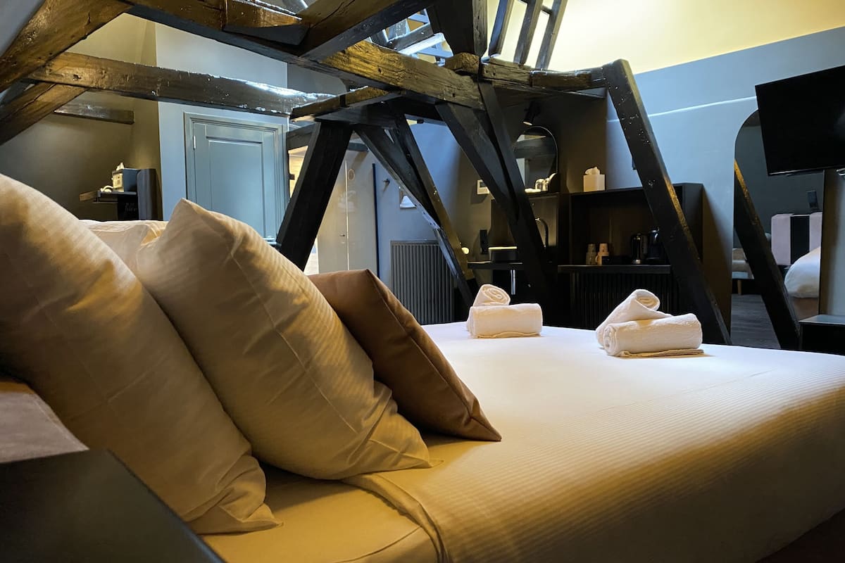 A hotel room with a bed, pillows, and towels. There is a wooden ceiling structure and a television on a stand.