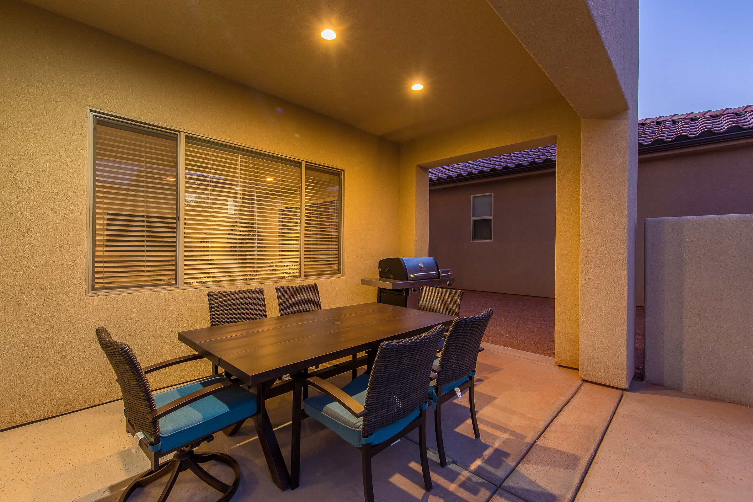 Um pátio externo com mesa de madeira e cadeiras de vime, churrasqueira e vista para uma casa ao fundo.