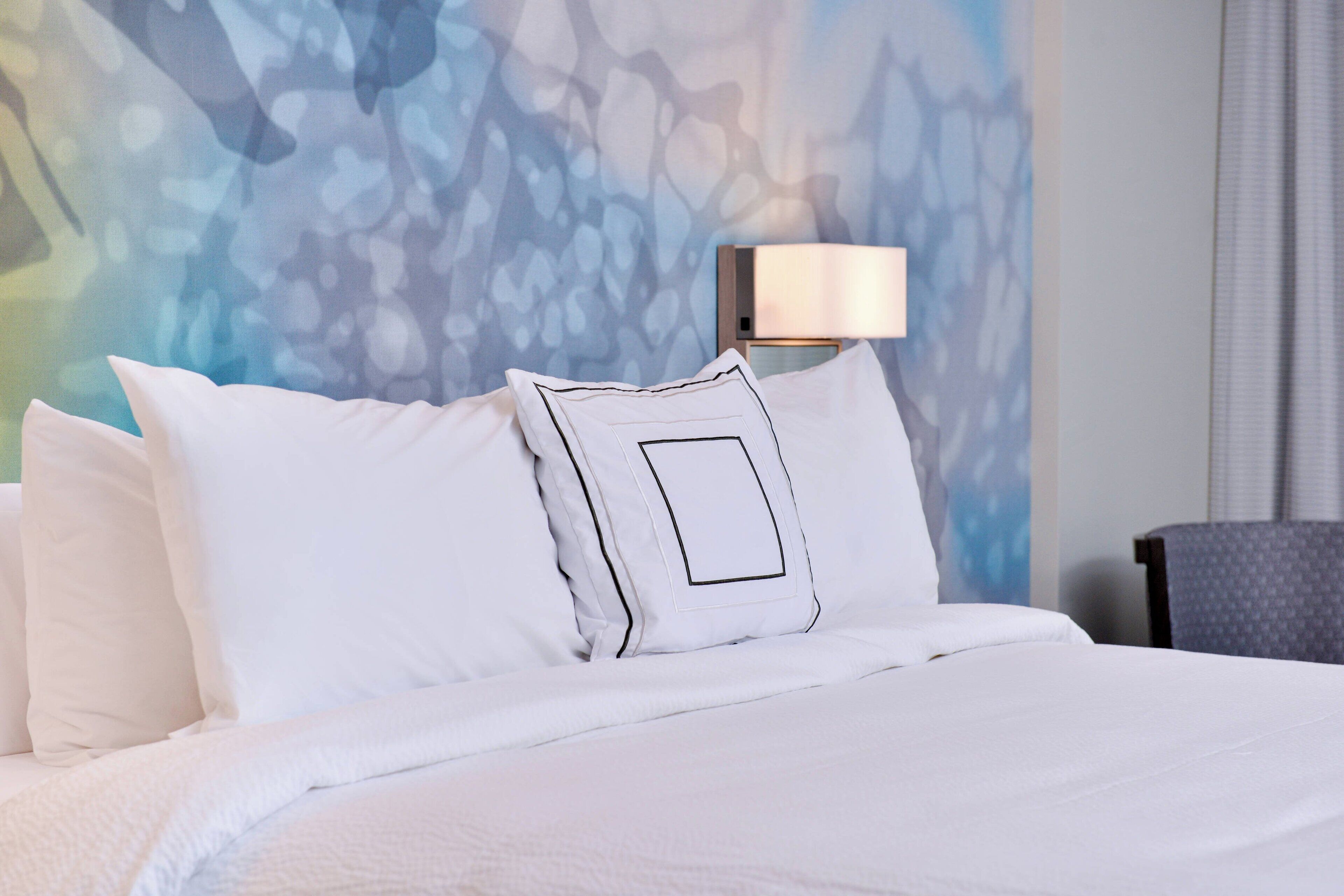 A neatly made bed with white linens and pillows, a modern lamp on the side, and a textured wall behind.