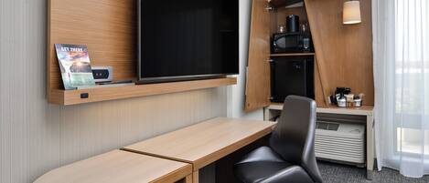 A hotel room with a wooden desk, a black office chair, a flat-screen TV mounted on the wall, a microwave, and a small kitchenette area.
