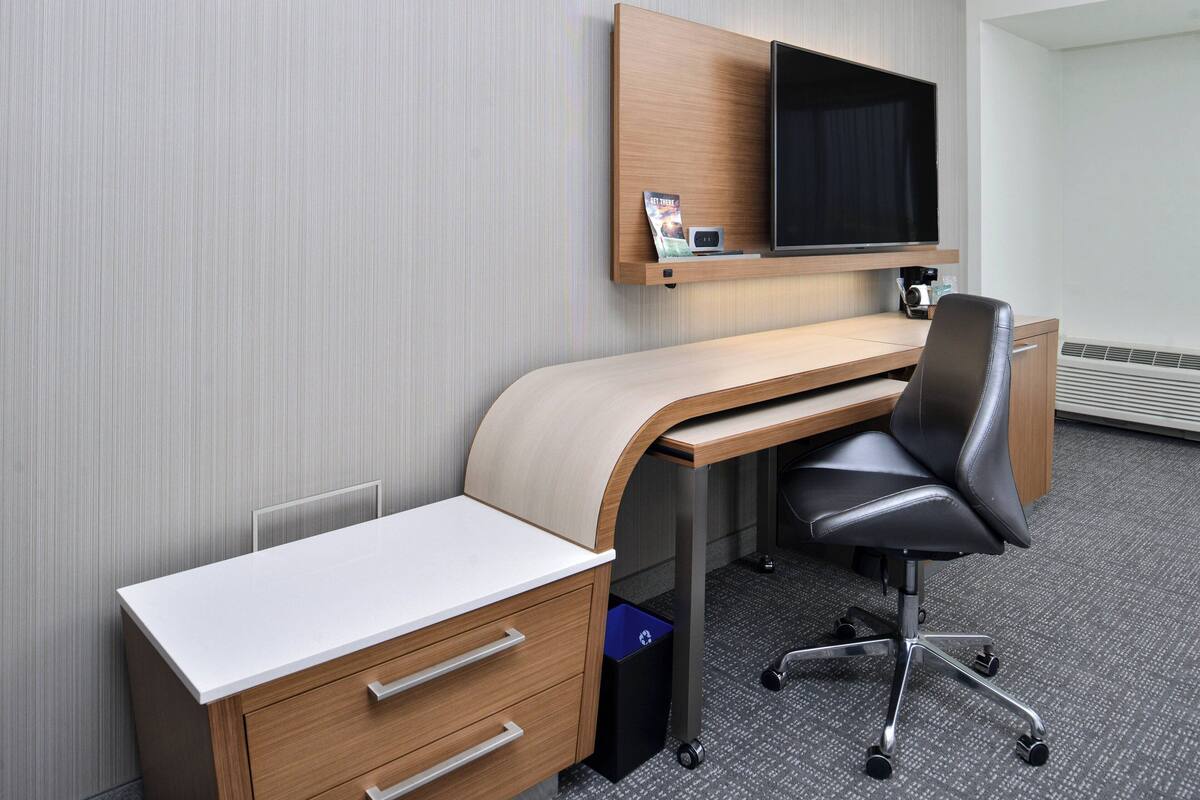 Una habitación de hotel con un escritorio de madera, una silla de oficina negra, un televisor de pantalla plana montado en la pared y un pequeño cubo de basura.