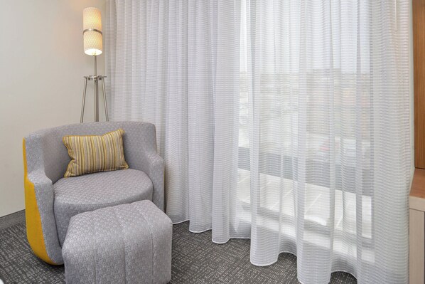 A modern hotel room with a grey sofa and ottoman, a floor lamp, sheer curtains, and a view of the outdoors.