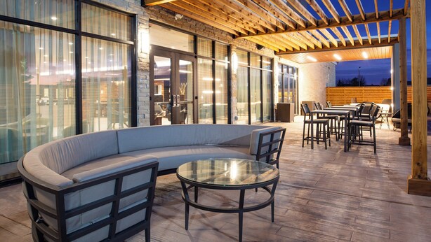 An outdoor patio with a curved sofa, round table, and chairs, wooden pergola, and large windows. - Courtyard by Marriott Kansas City Olathe (Olathe)
