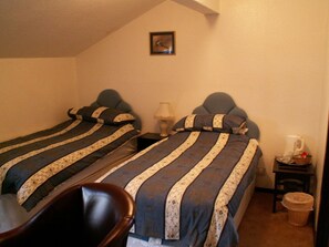 A small, cozy bedroom with two single beds, a nightstand with a lamp, a chair, and a small table with a basket and a kettle.