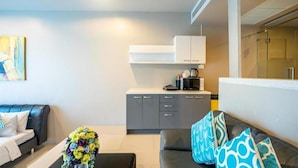 A modern living room with a black sofa, a coffee table with a floral arrangement, and a grey kitchen area with cabinets and a countertop. - The Ark By Veloche Group (Karon)