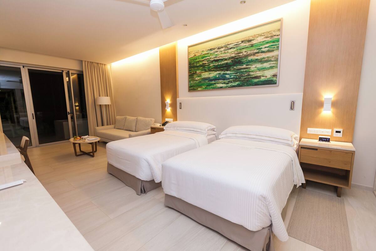 Ein Hotelzimmer mit zwei Betten, einem Sofa, einem Schreibtisch und einer Glasschiebetür, die auf einen Balkon führt.