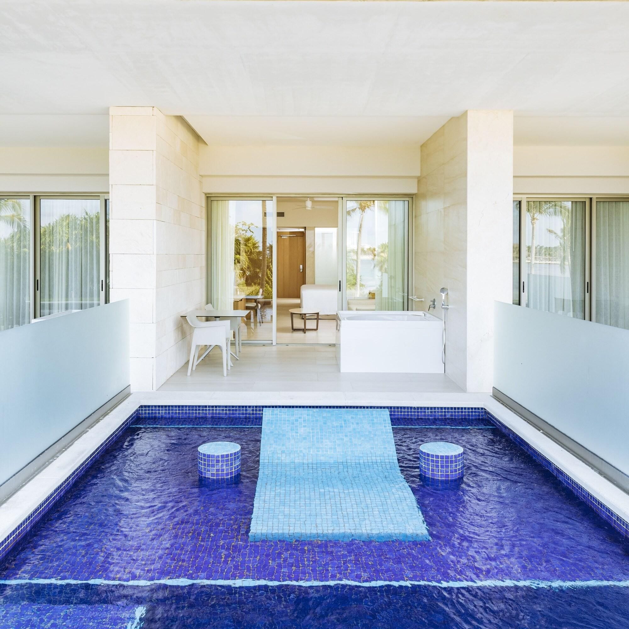 A modern indoor swimming pool with a blue mosaic floor, a seating area with a table and chairs, and a large bathtub.