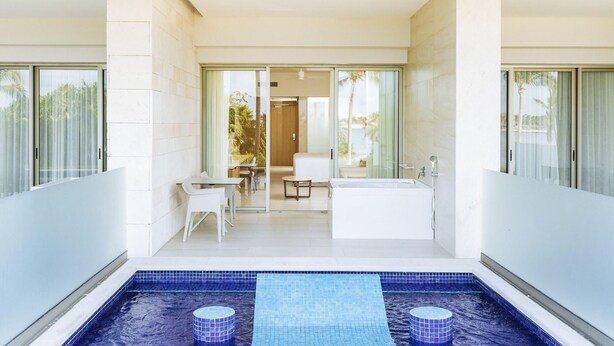 A modern indoor swimming pool with a blue mosaic floor, a seating area with a table and chairs, and a large bathtub.