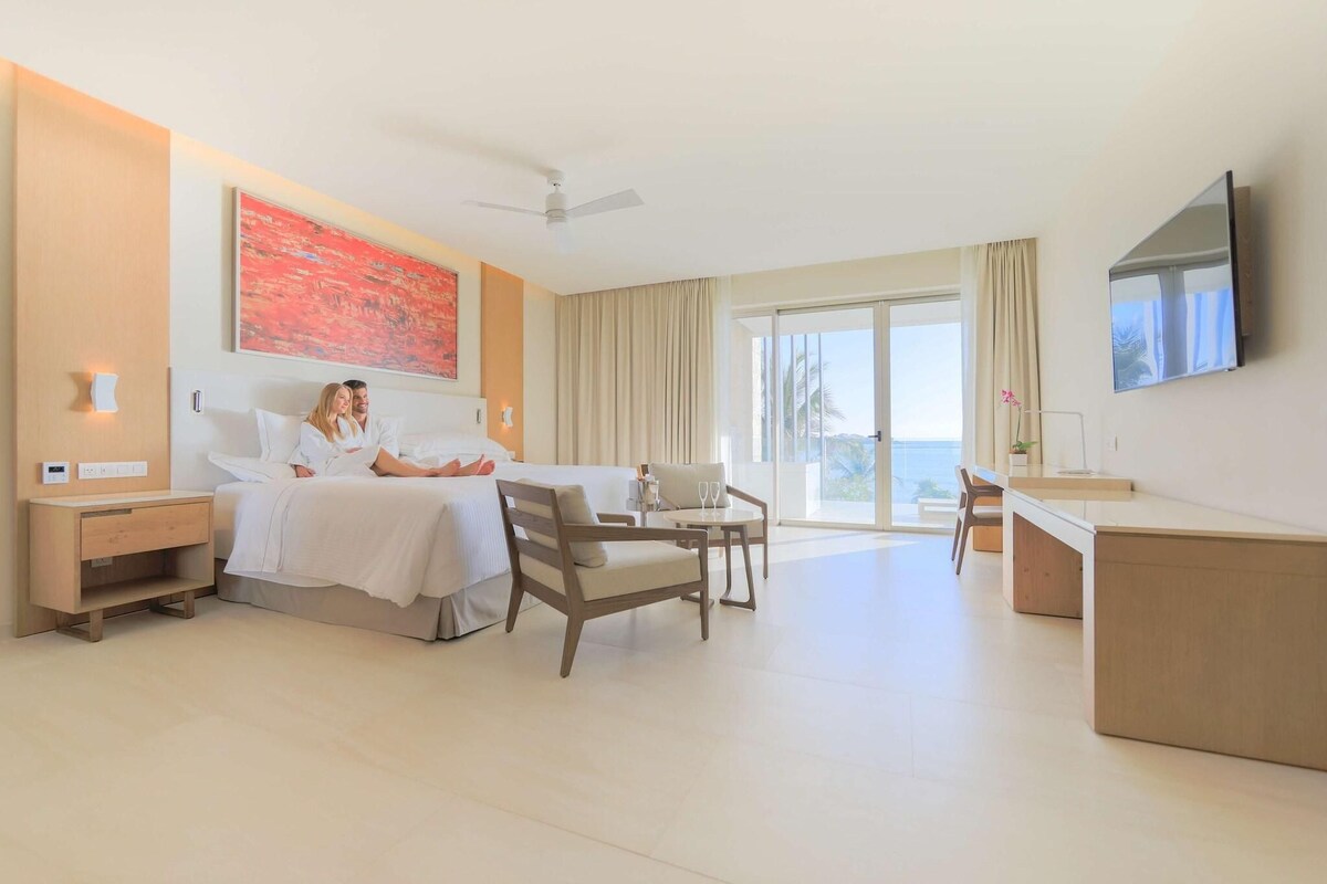 Ein Hotelzimmer mit Bett, Nachttisch, Stuhl, Schreibtisch und einem großen Fenster mit Vorhängen.