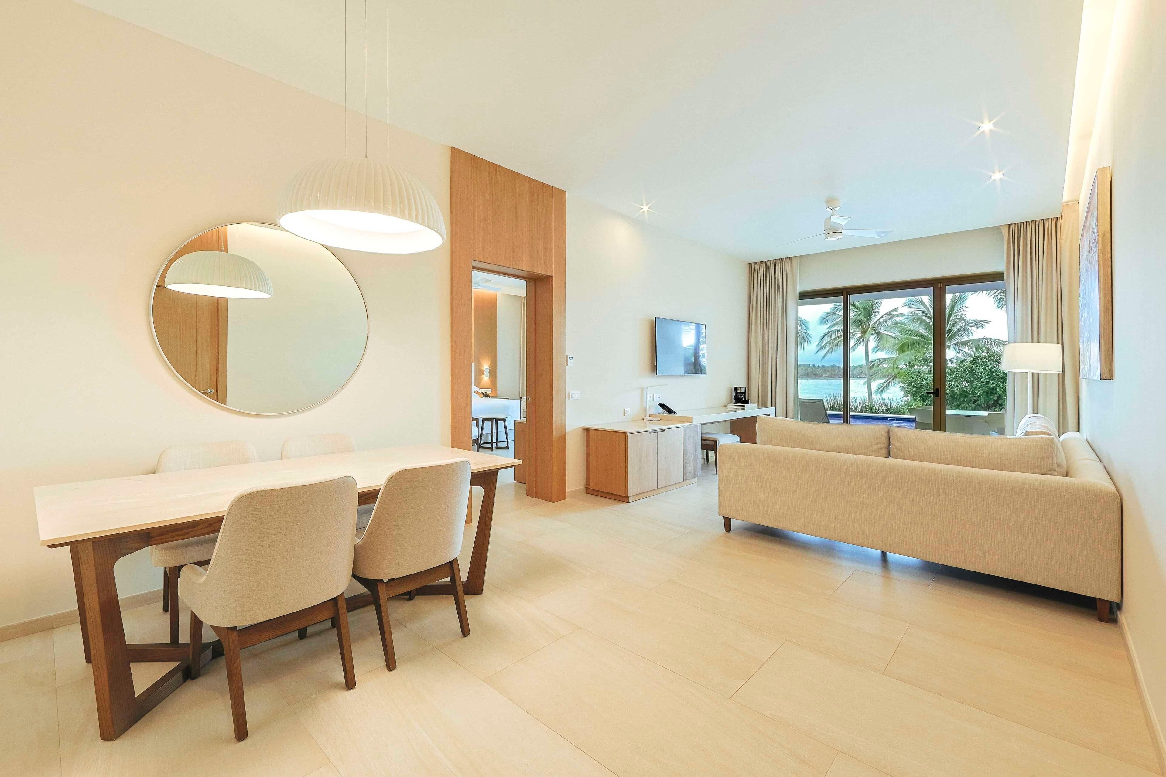 A modern hotel room with a dining area, a sofa, a TV, and large windows with a view of palm trees.
