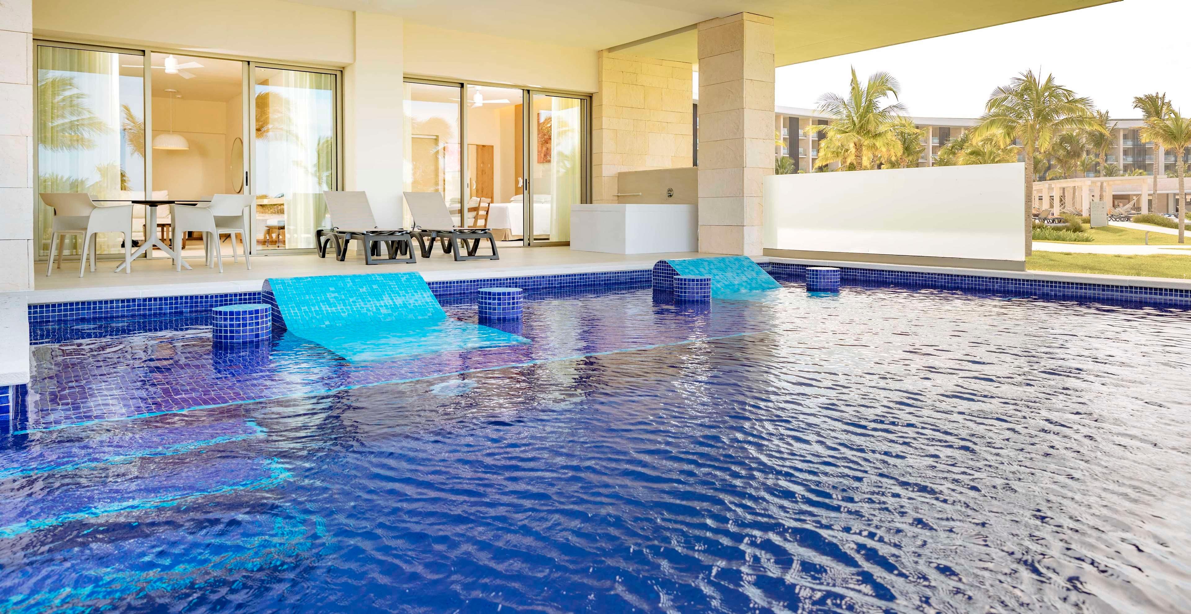 Una piscina interior moderna con agua azul cristalina, un área acristalada con mesa y sillas, y vistas a palmeras y edificios al exterior.