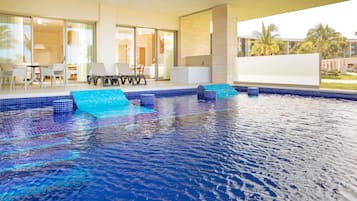 Una piscina interior moderna con agua azul cristalina, un área acristalada con mesa y sillas, y vistas a palmeras y edificios al exterior.