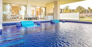 A modern indoor swimming pool with clear blue water, a glass-enclosed area with a table and chairs, and a view of palm trees and buildings outside. - Barcelo Maya Riviera (Xpu-Ha)