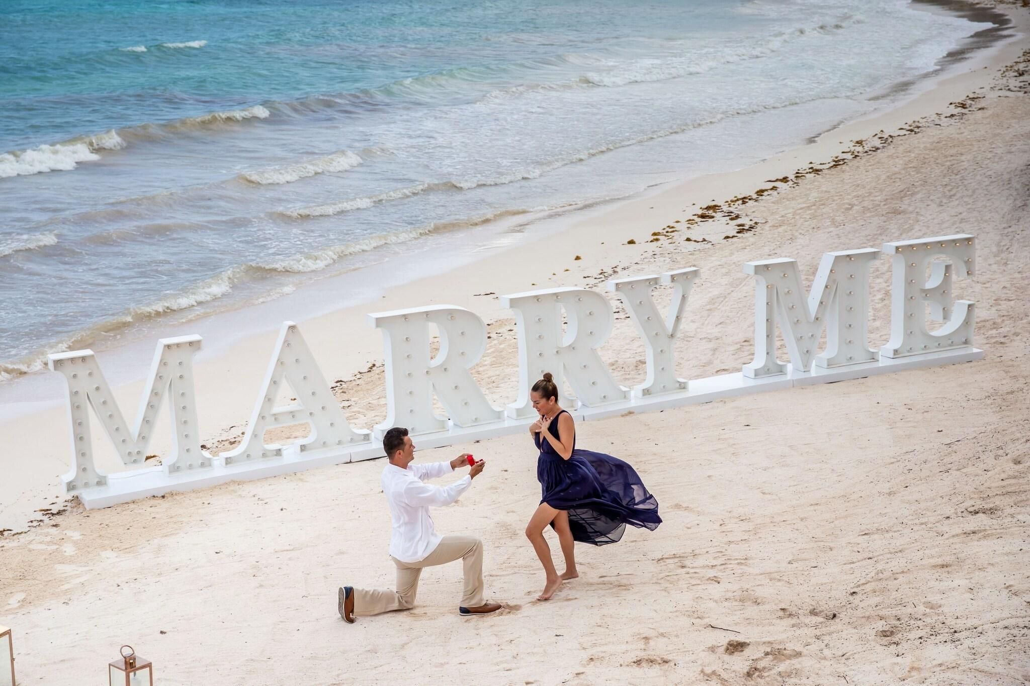 A beach setting with a large "MARRY ME" sign and a man proposing to a woman.