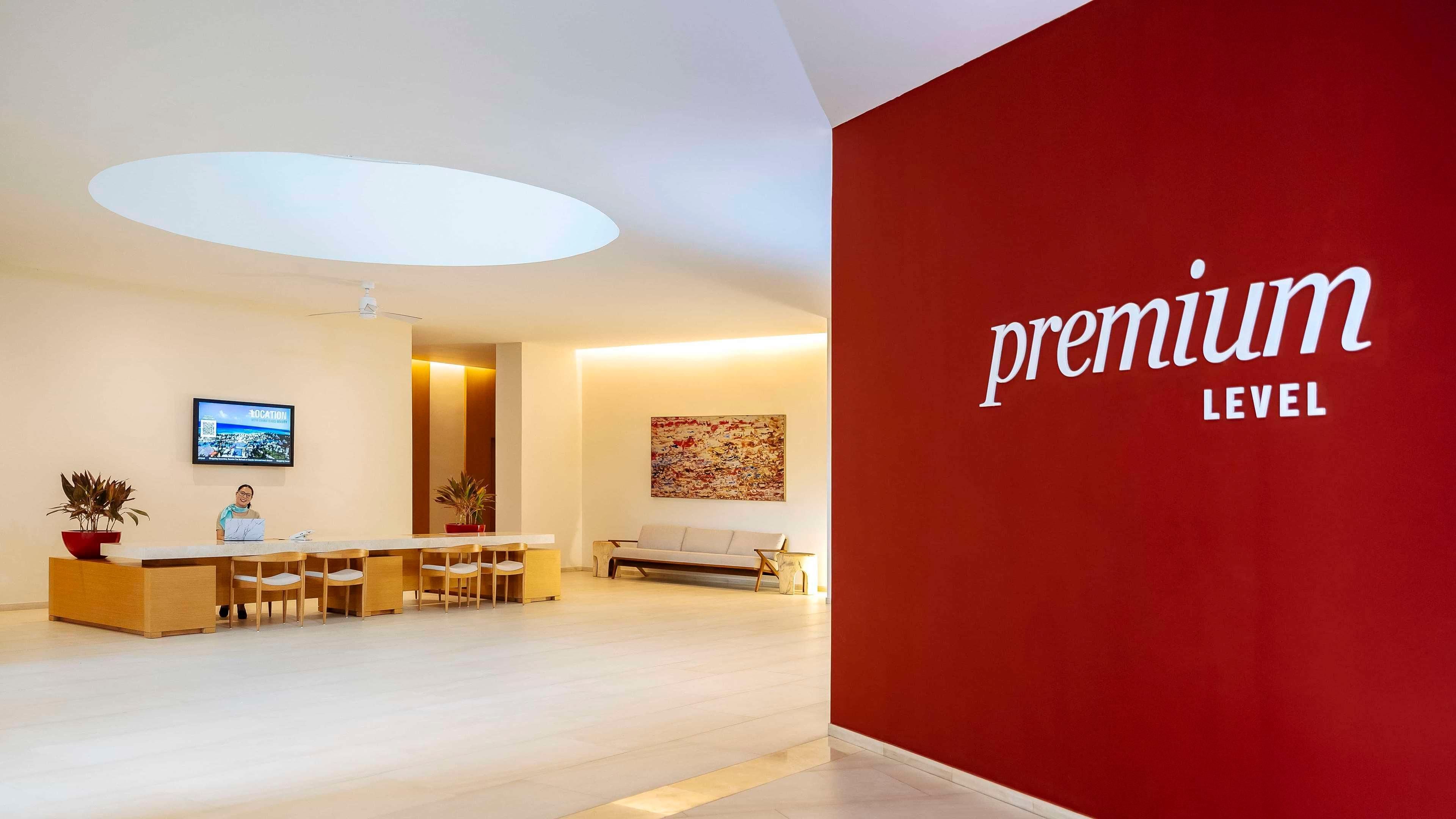 Zona de recepción de hotel con pared roja que muestra el texto "premium level", un mostrador de recepción, una zona de estar con sofá y una pantalla de televisión en la pared.