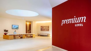 A hotel reception area with a red wall displaying "premium level" text, a reception desk, a seating area with a sofa, and a TV screen on the wall. - Barcelo Maya Riviera (Xpu-Ha)