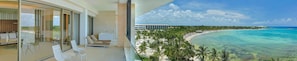 A balcony with a glass door leading to a room with a sofa and chairs, overlooking a beach and ocean. - Barcelo Maya Riviera (Xpu-Ha)