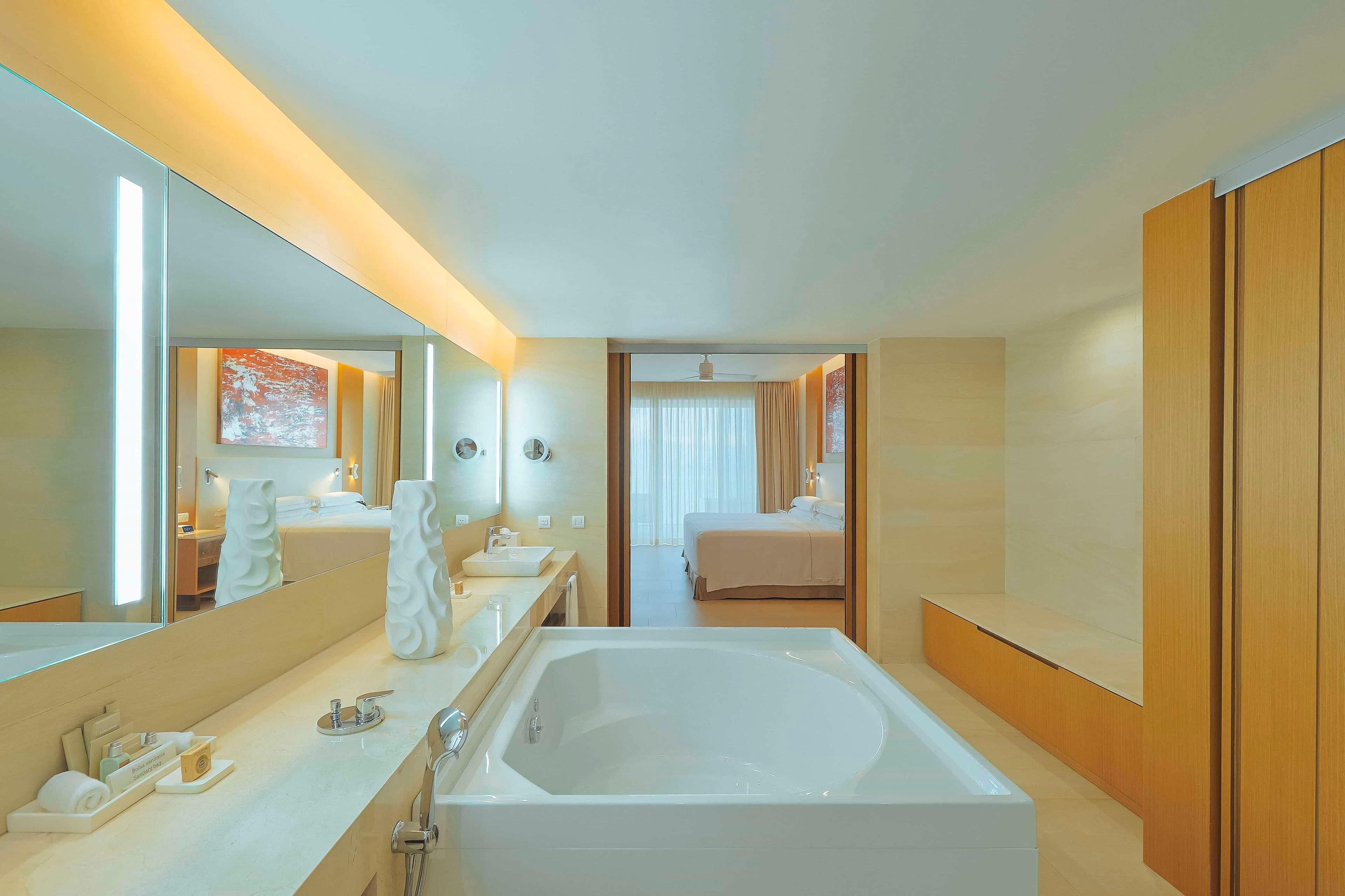 Un moderno baño de hotel con una bañera grande, un tocador con espejo y una habitación separada visible a través de una puerta abierta.