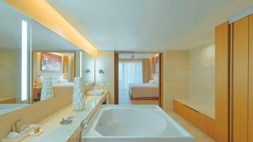 Un moderno baño de hotel con una bañera grande, un tocador con espejo y una habitación separada visible a través de una puerta abierta.