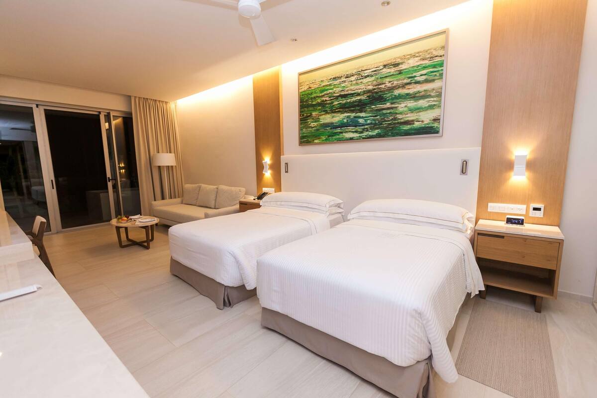 Ein Hotelzimmer mit zwei Betten, einer Schlafcouch, einem Schreibtisch und einem großen Fenster.