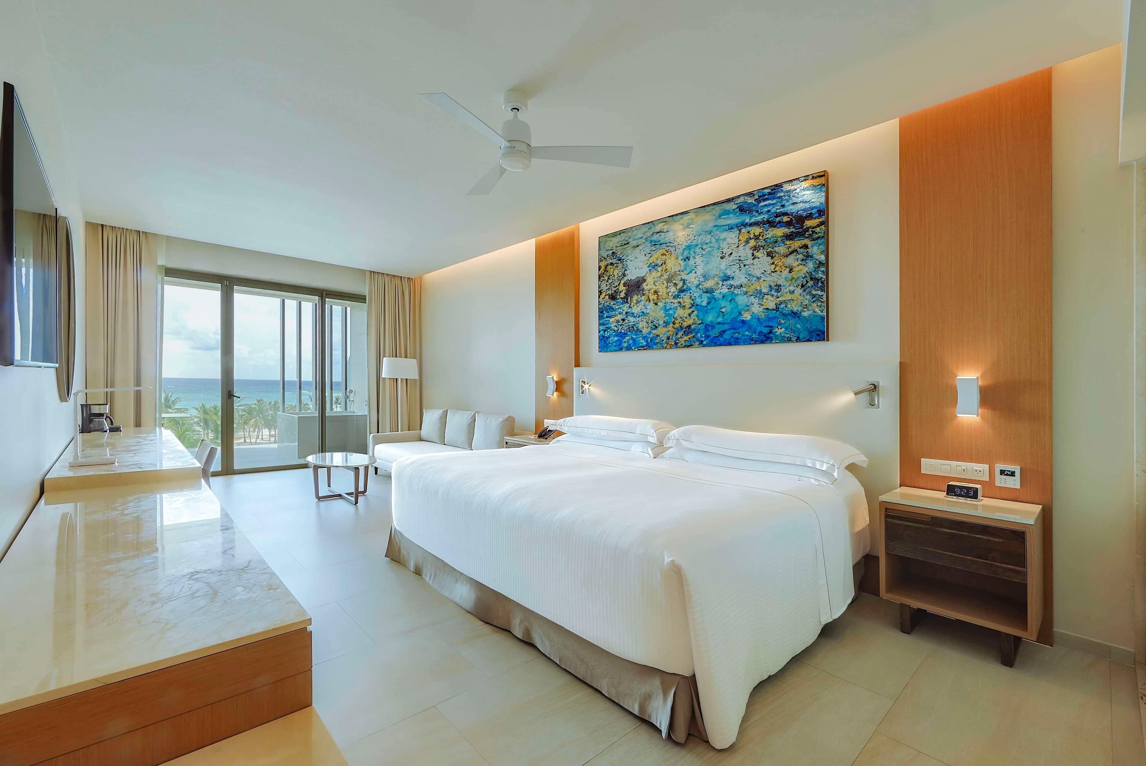 A modern hotel room with a large bed, a desk, a flat-screen TV, a ceiling fan, a painting, and a view of the ocean.
